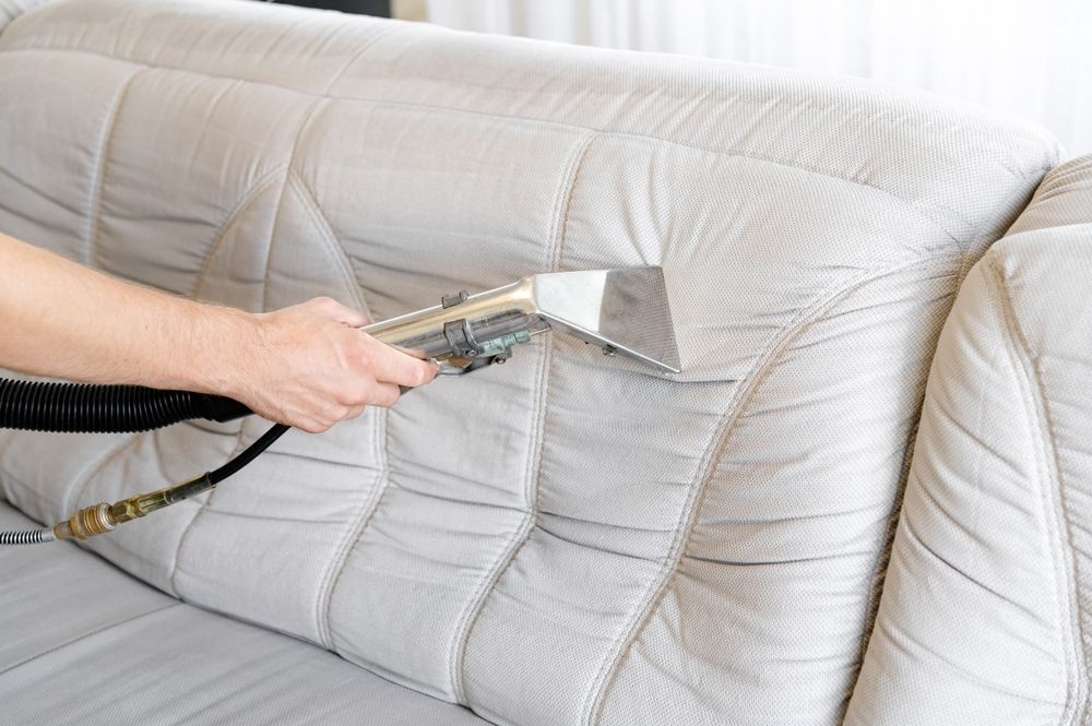 Person cleaning a light-colored sofa with a handheld upholstery cleaner.