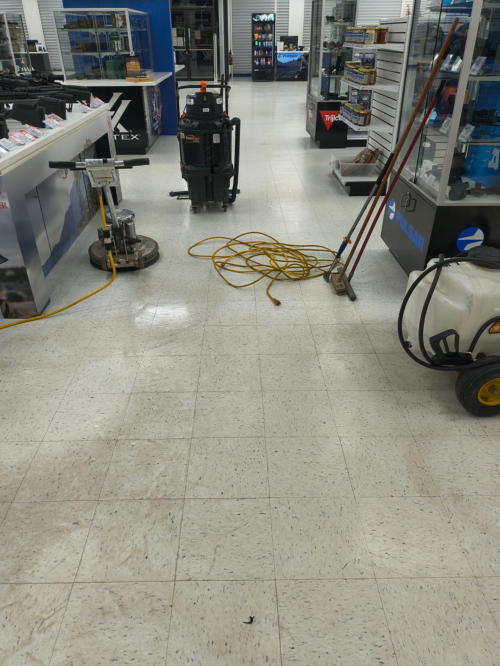 Inside a store, cleaning equipment on floor: floor buffer, spray tank, and electrical cords. Shelves and merchandise visible.