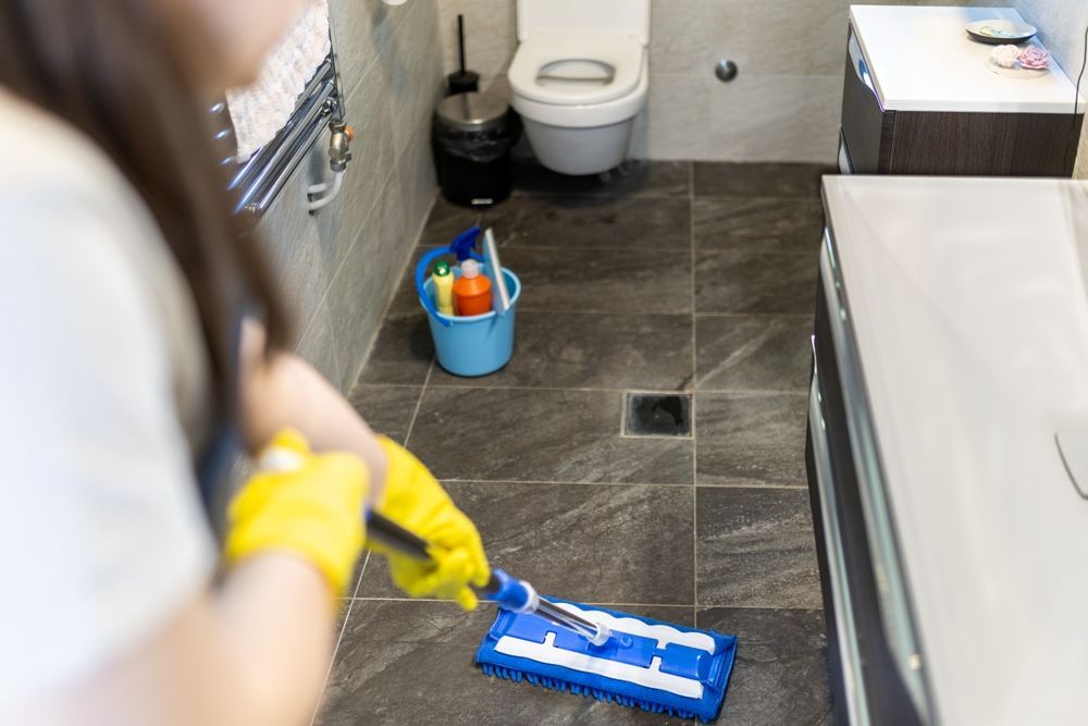 Person mopping a tiled bathroom floor, wearing yellow gloves. A blue bucket with cleaning supplies sits nearby.