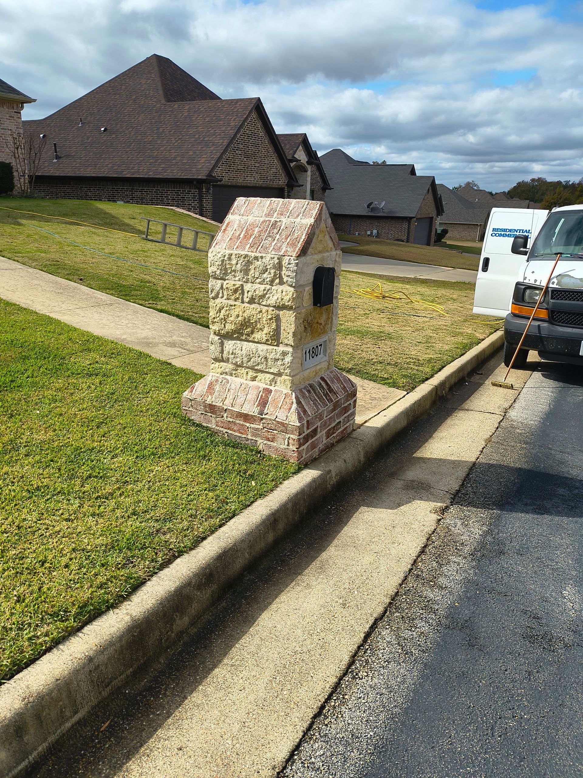 Stone mailbox with house-shaped top, set on grass next to a sidewalk and a street, in front of a row of houses.