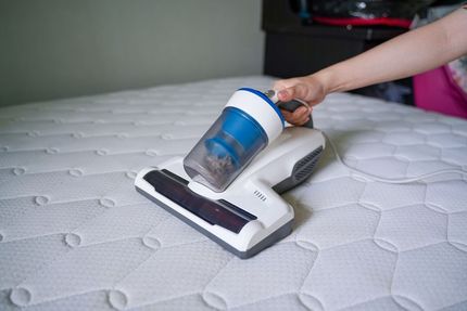 Hand using a small vacuum to clean a white mattress.