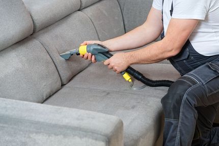 Person cleaning a gray couch with a handheld upholstery cleaner.
