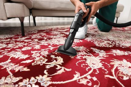 Person using a carpet cleaner on a red patterned rug near a beige sofa.