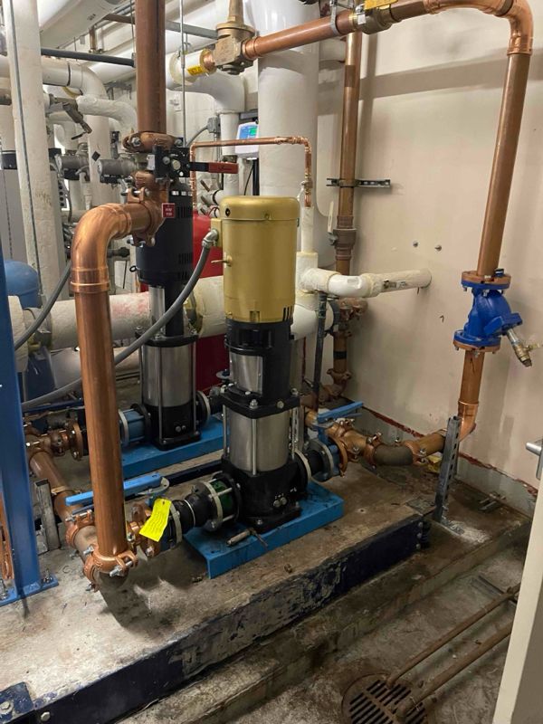 Two water pumps with copper pipes in an industrial setting. Yellow and black components, blue metal base, and white wall.