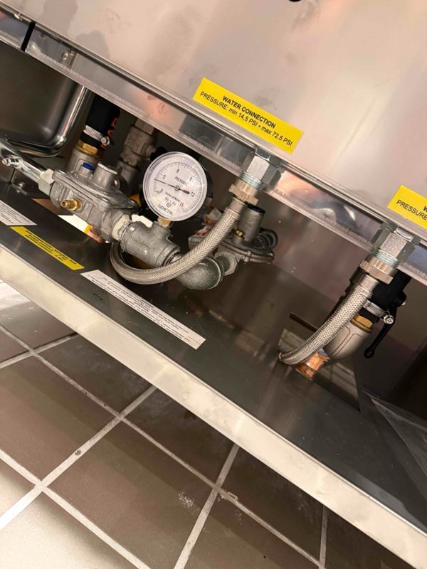 Plumbing components inside a stainless steel compartment, including a pressure gauge and water connections.