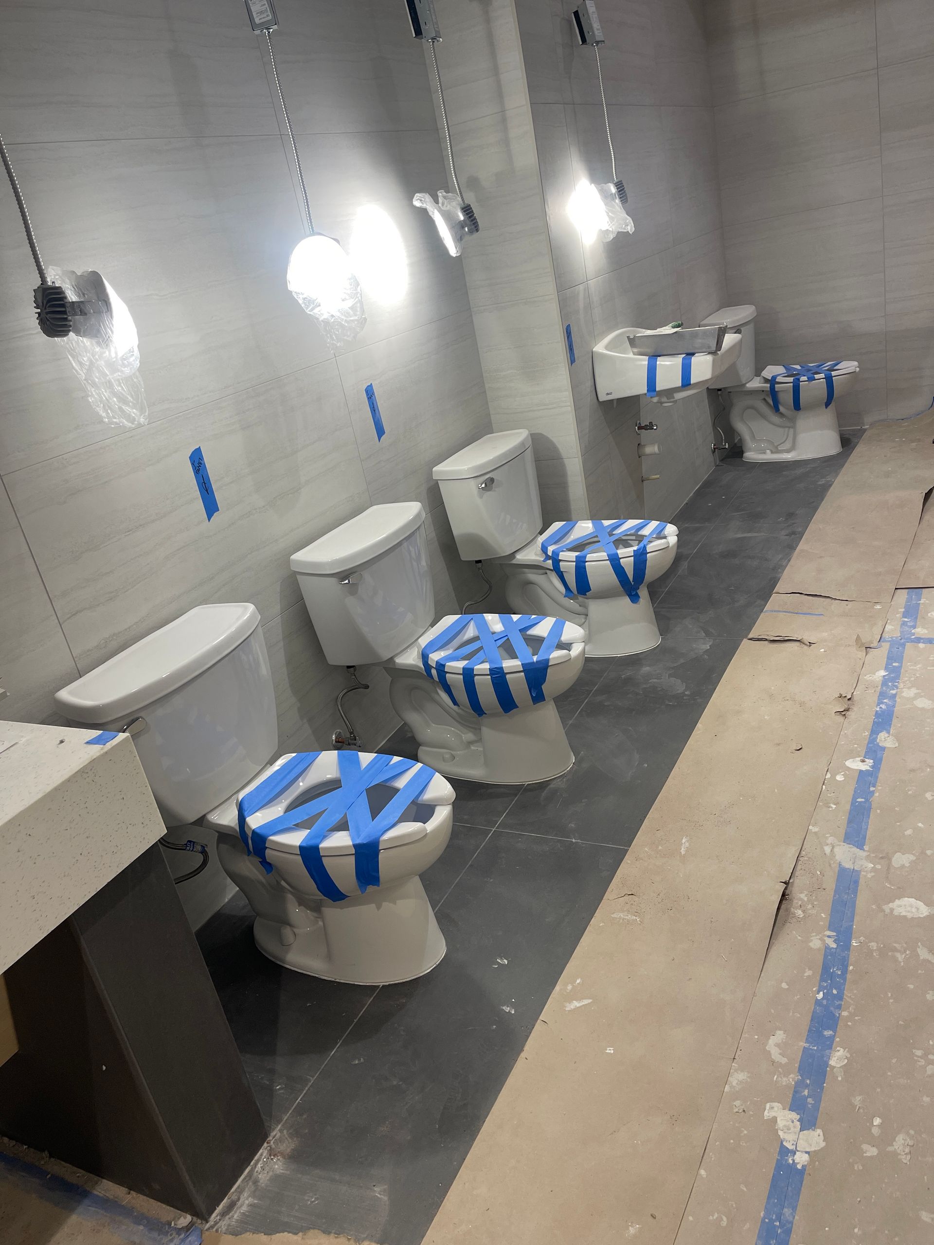 Multiple toilets in a row, taped, under construction lights in a gray-walled room.