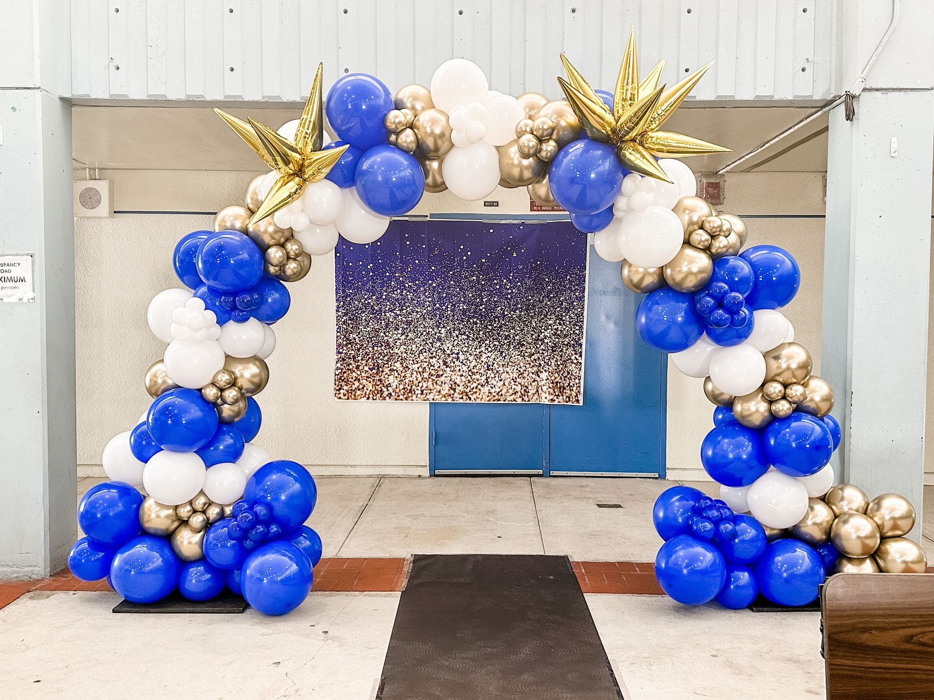 A blue , white and gold balloon arch is sitting in front of a building.