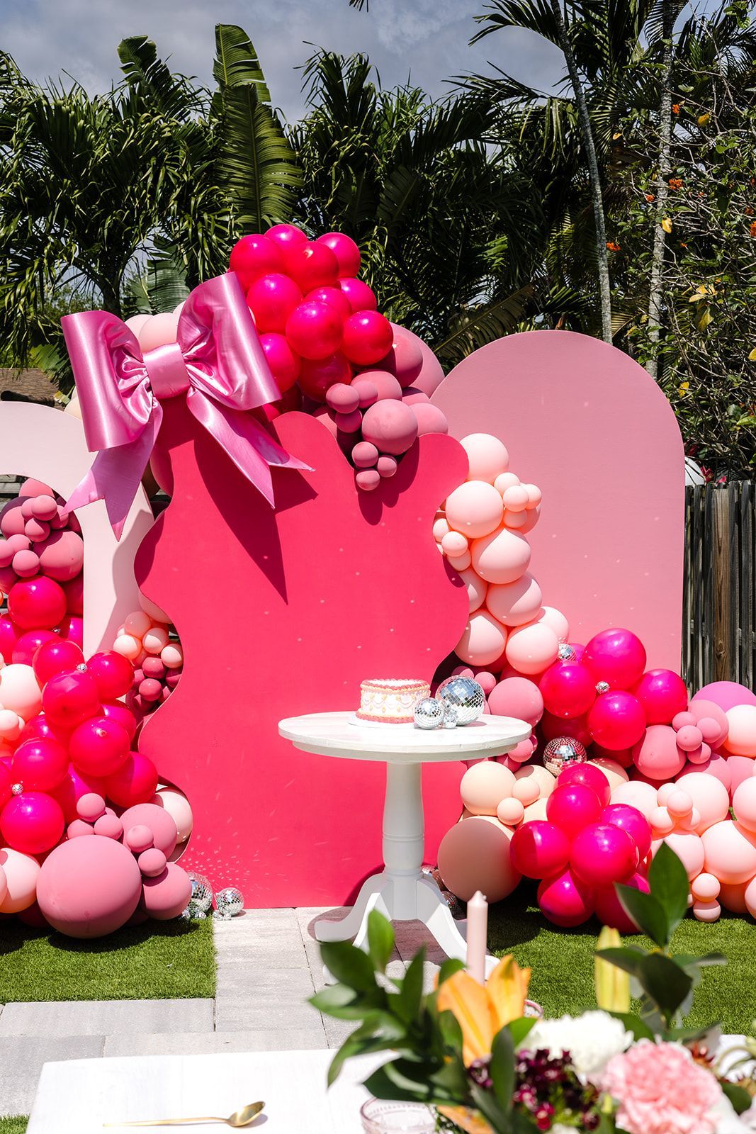 A table with a cake on it is surrounded by pink balloons and flowers.
