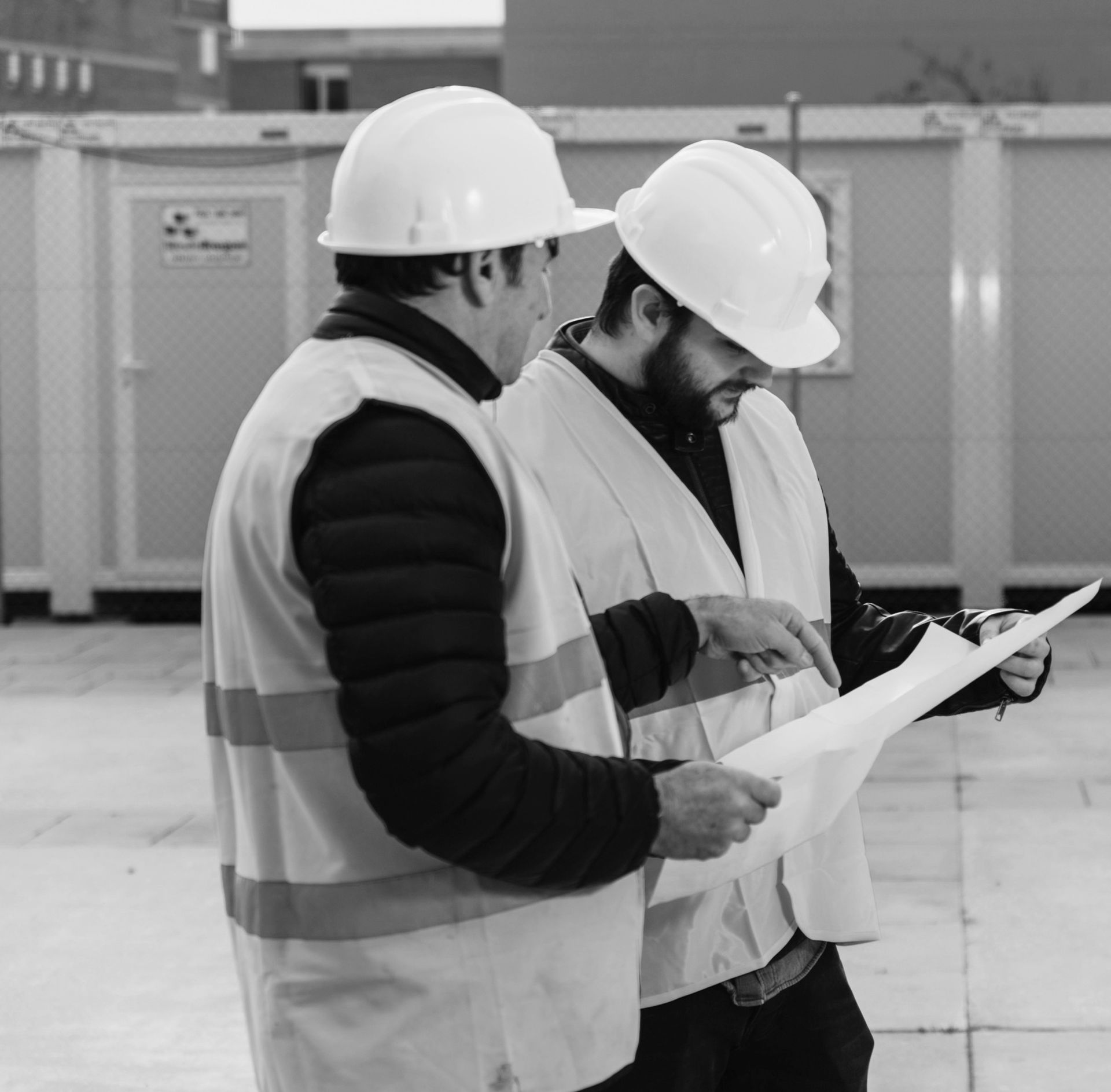Gaining pattern intelligence through operational similarity. Two construction supervisors wearing white hard hats and high-visibility safety vests conferring over a site blueprint, illustrating how group captives align members based on shared operational methods and safety protocols.