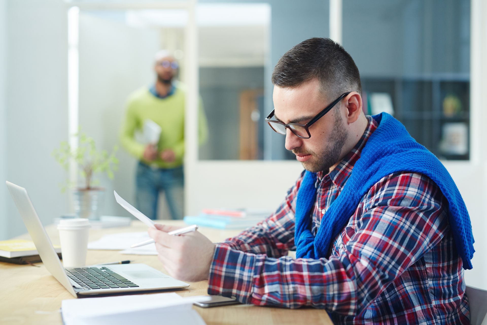 Accountability through intentional risk behavior and documentation. A focused professional in a plaid shirt and glasses sits at a desk, carefully reviewing a physical document while working on a laptop, representing the financial maturity and detailed documentation required in group captive insurance.