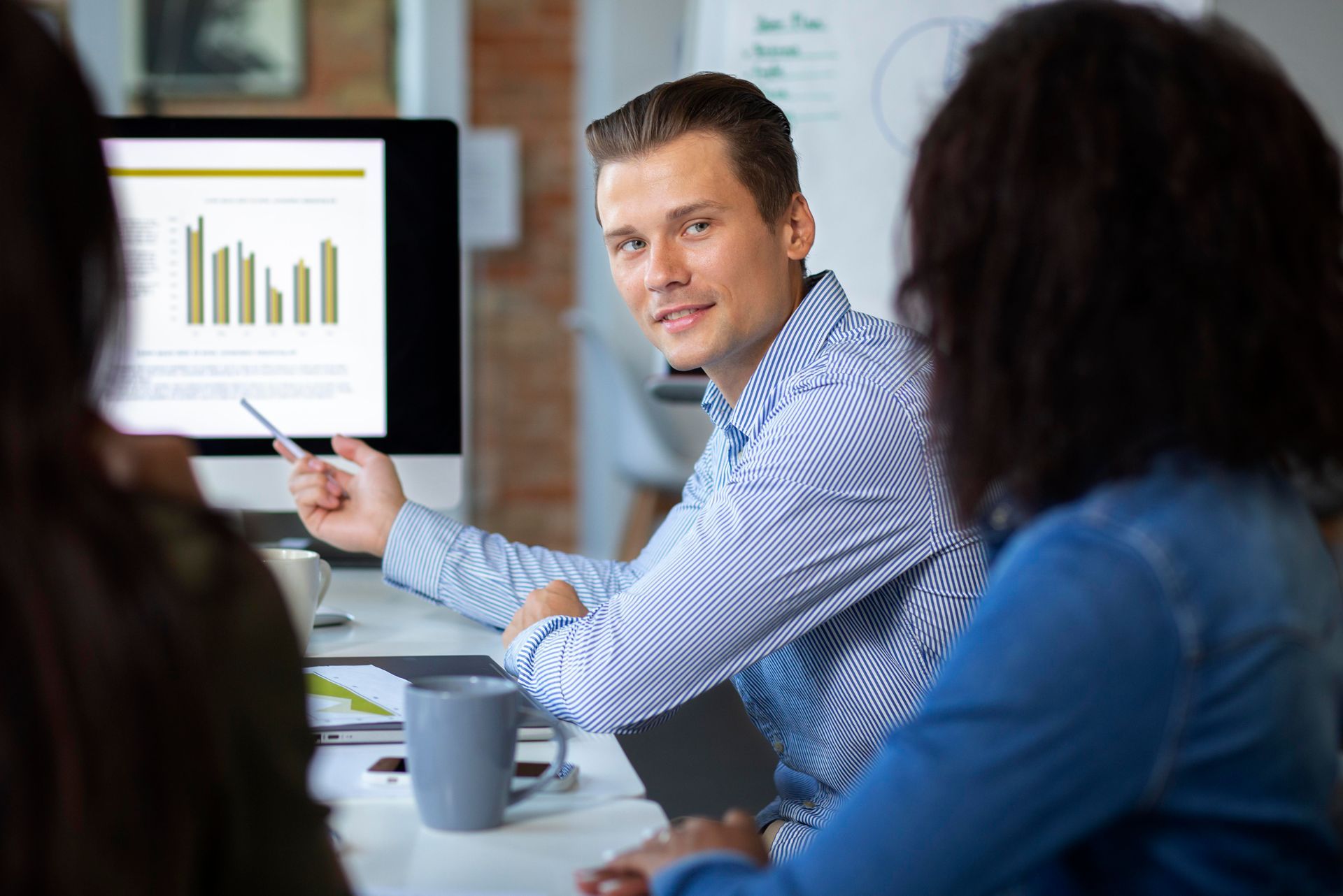Converting safety performance into a financial advantage. A business professional in a striped shirt pointing at a data chart on a monitor while engaged in a collaborative discussion with colleagues, symbolizing the economic shift toward group captive insurance models.