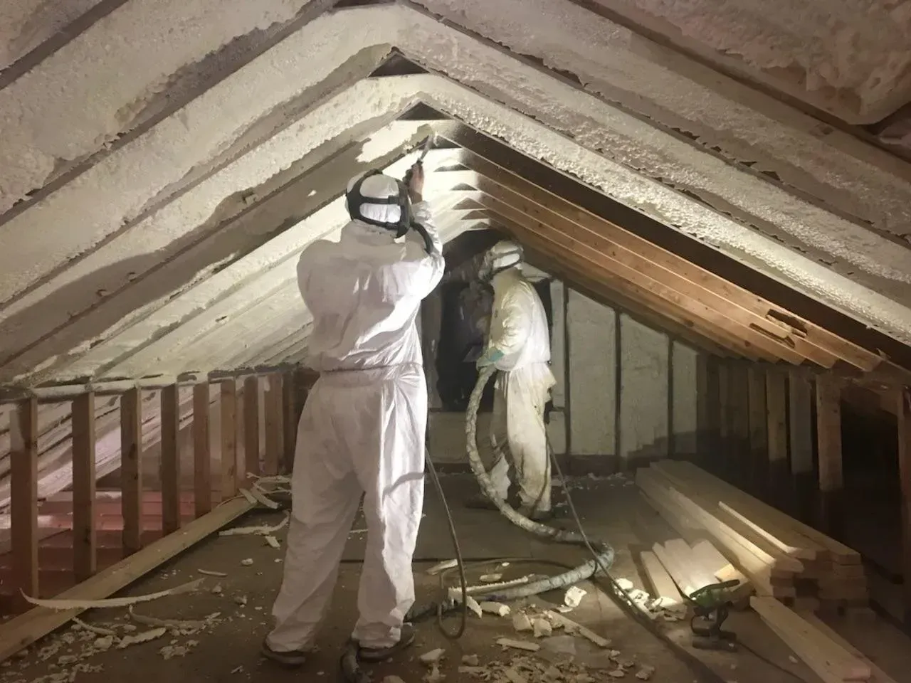 A man in a white suit is spraying foam in an attic.