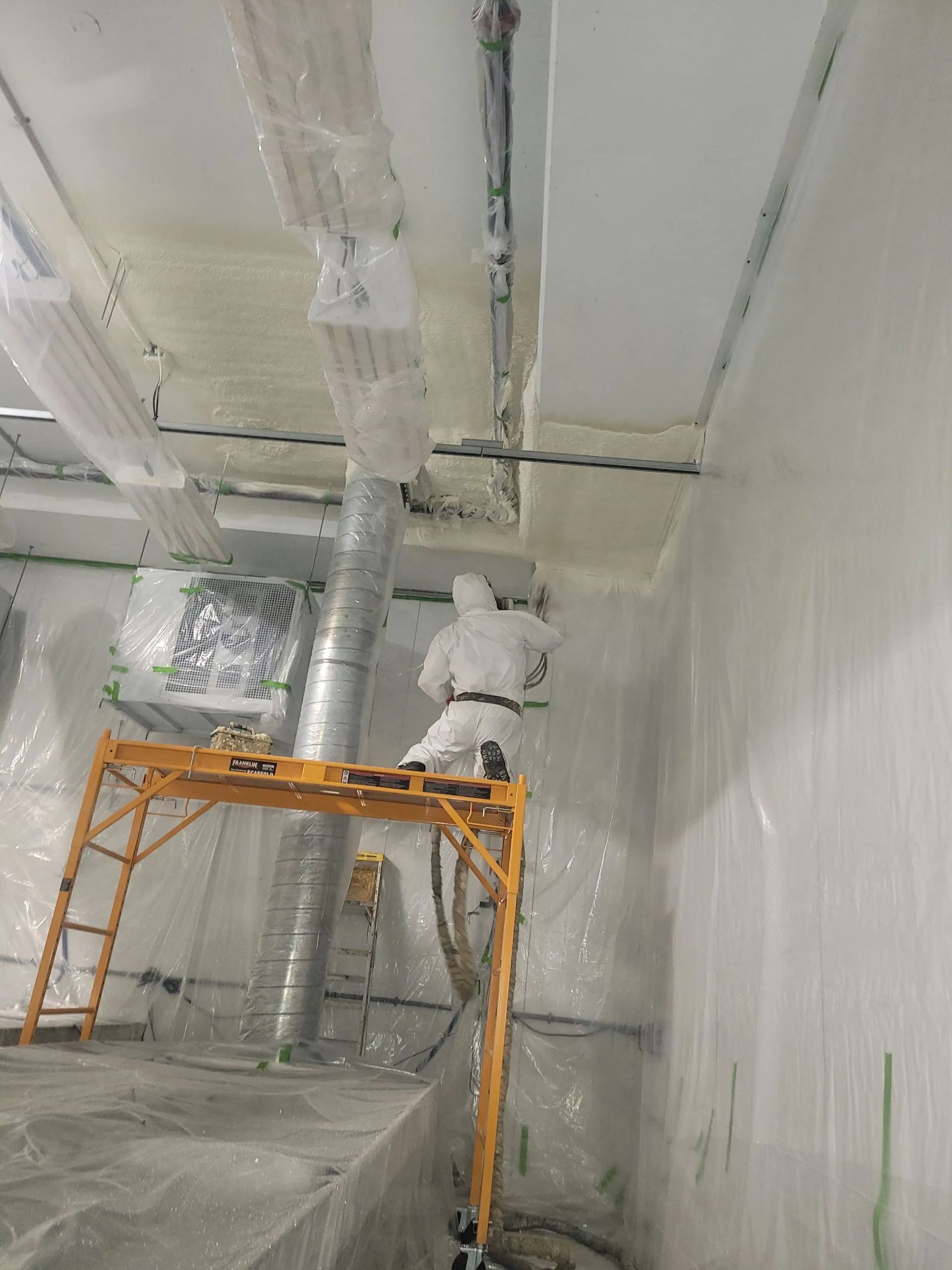 Worker on an orange scaffold painting white ceiling pipes in a covered industrial room.
