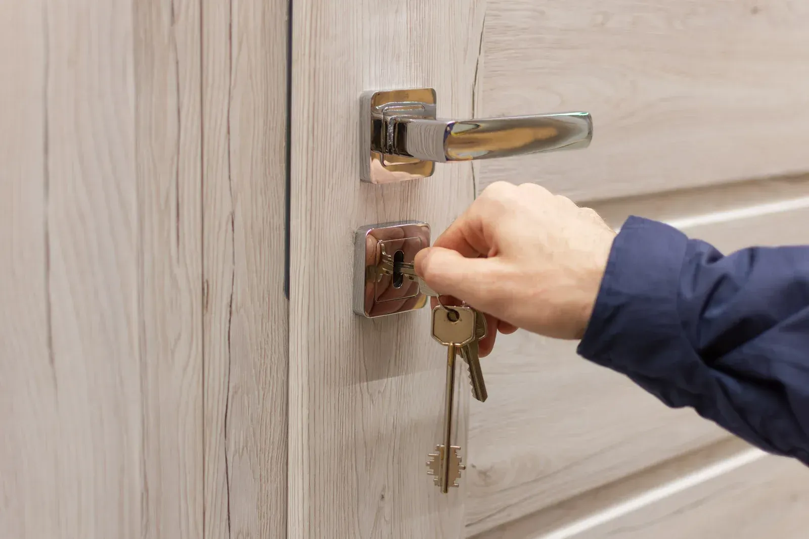 Hand inserting key into a light-colored wooden door's lock with a chrome handle.
