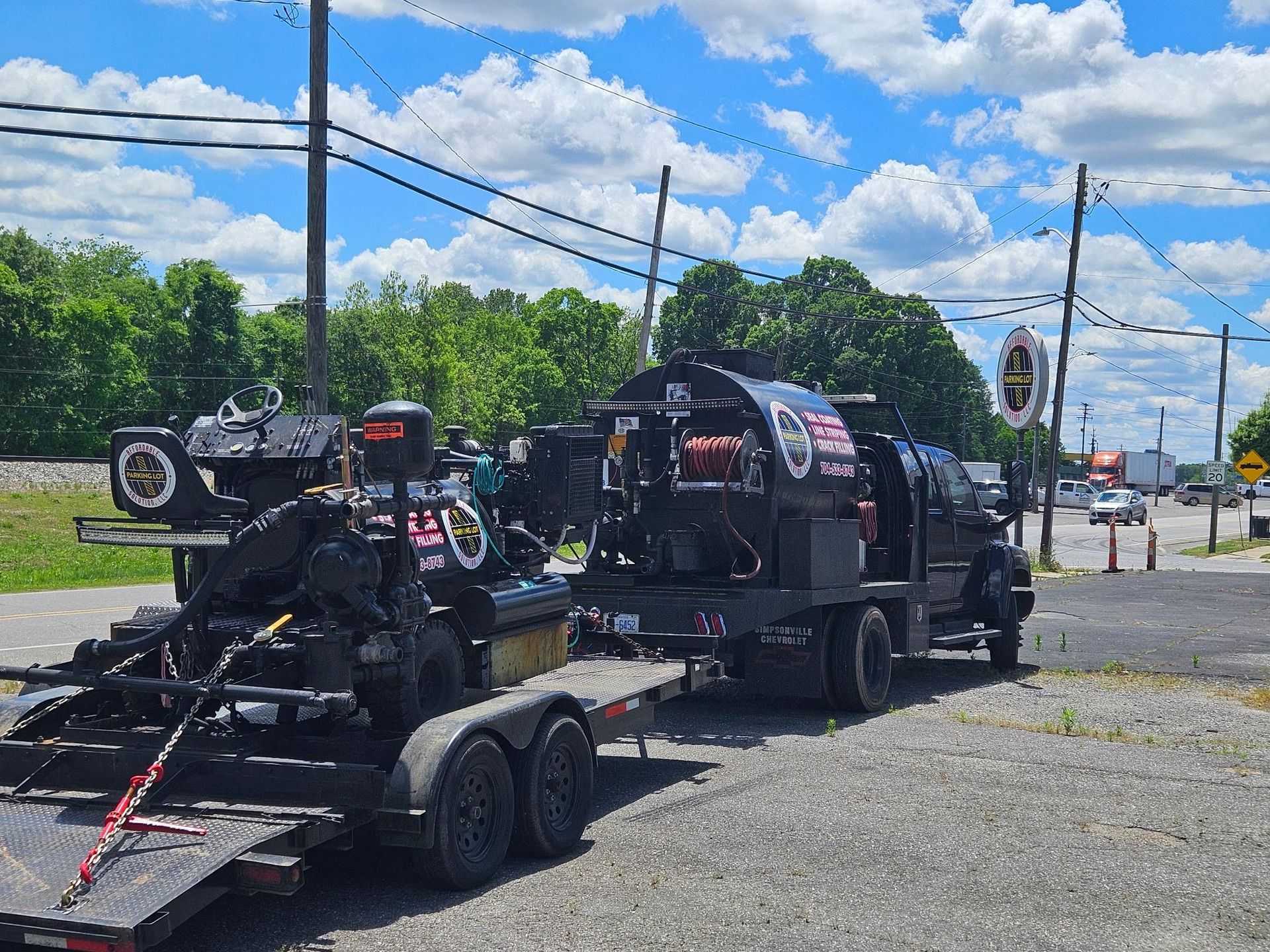 A black truck towing a flatbed trailer loaded with specialized black construction or maintenance equipment outdoors.