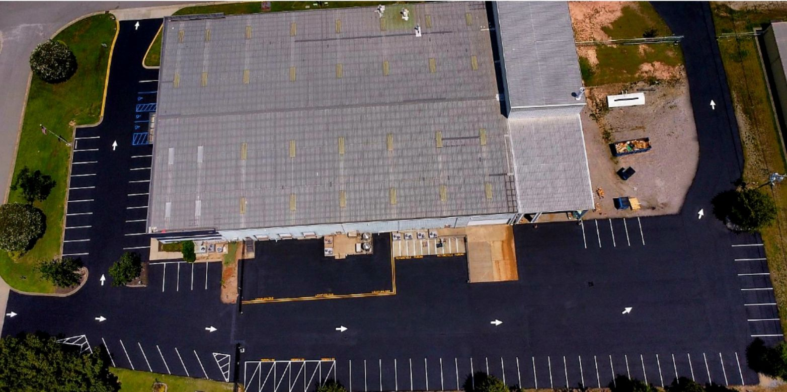 An aerial view of a rectangular warehouse building surrounded by a newly paved asphalt parking lot with white marked spaces.