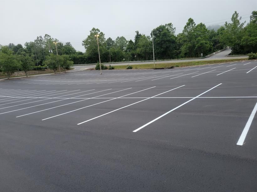 An empty, freshly paved asphalt parking lot with white painted lines and surrounding green trees under a cloudy sky.