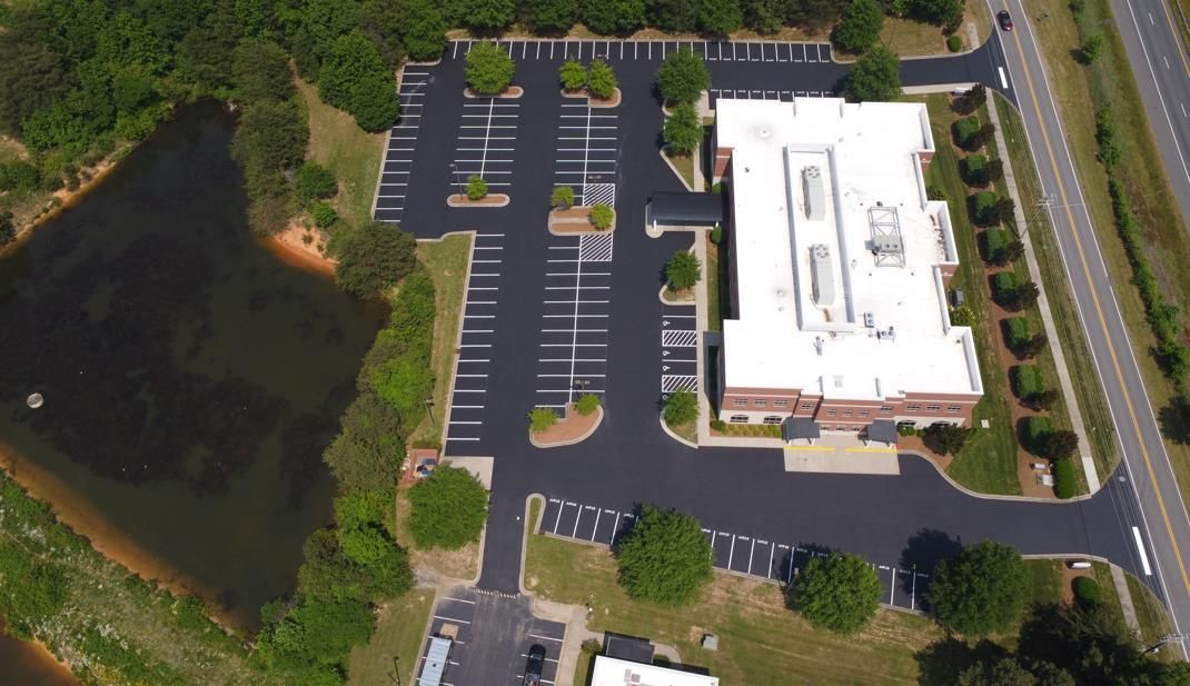 An empty, freshly paved asphalt parking lot with white painted lines and surrounding green trees under a cloudy sky.