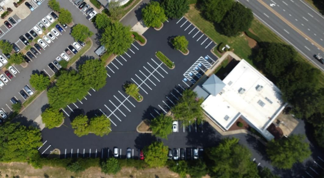 An empty, freshly paved asphalt parking lot with white painted lines and surrounding green trees under a cloudy sky.