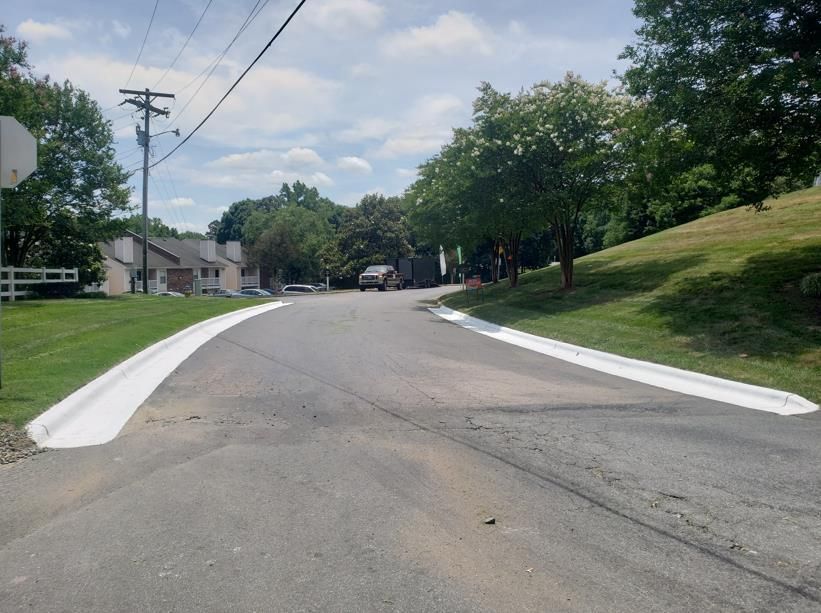 An empty, freshly paved asphalt parking lot with white painted lines and surrounding green trees under a cloudy sky.