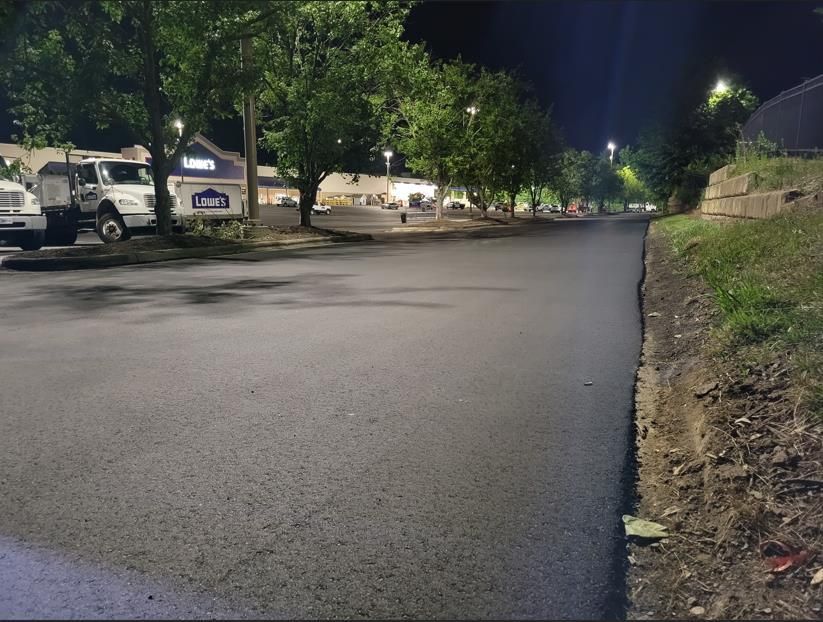 A freshly paved asphalt road at night, with a Lowe's store and parking lot illuminated in the background.