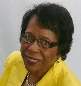 A person with dark hair and glasses wears a bright yellow jacket and a pearl necklace, smiling against a white background.