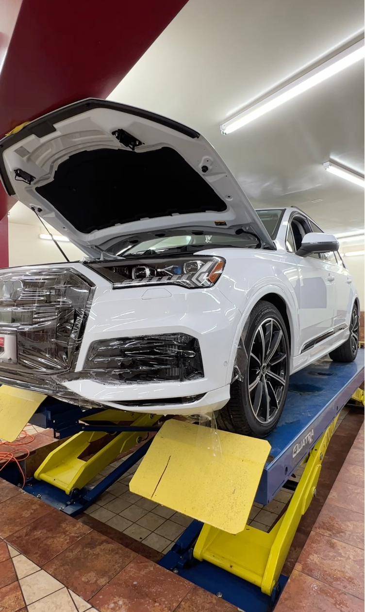 A white car is sitting on a lift with its hood open  while getting a tint installation service.
