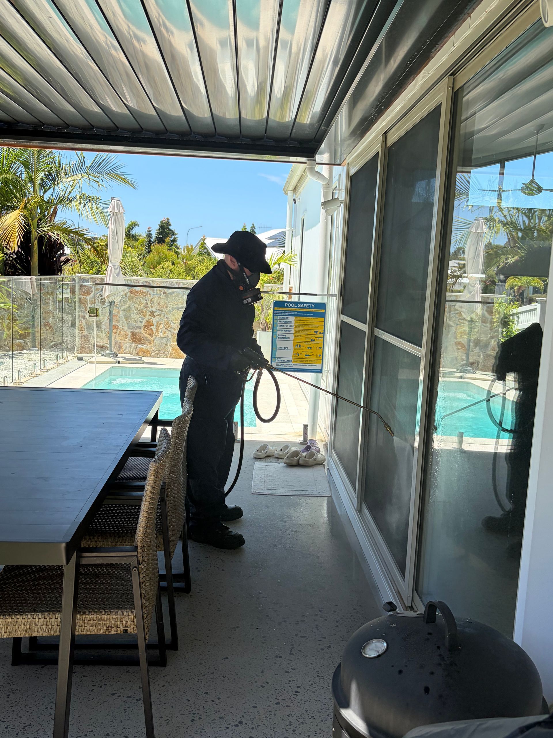 A pest control technician in uniform sprays the edges of a sliding glass door on a patio overlooking a swimming pool  — FUMATEX In Port Macquarie, NSW