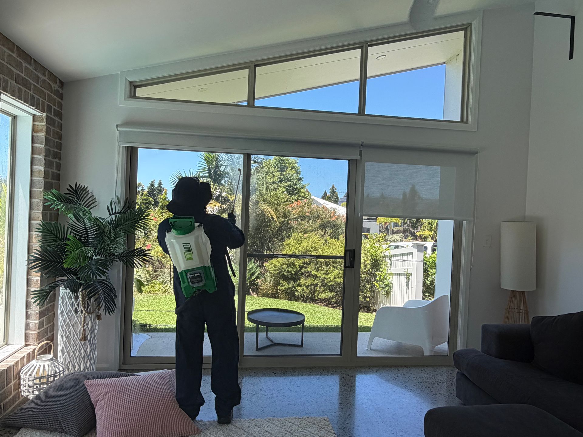 A person wearing a backpack sprayer treats a large glass door in a sunlit modern living room with a patio view  — FUMATEX In Port Macquarie, NSW