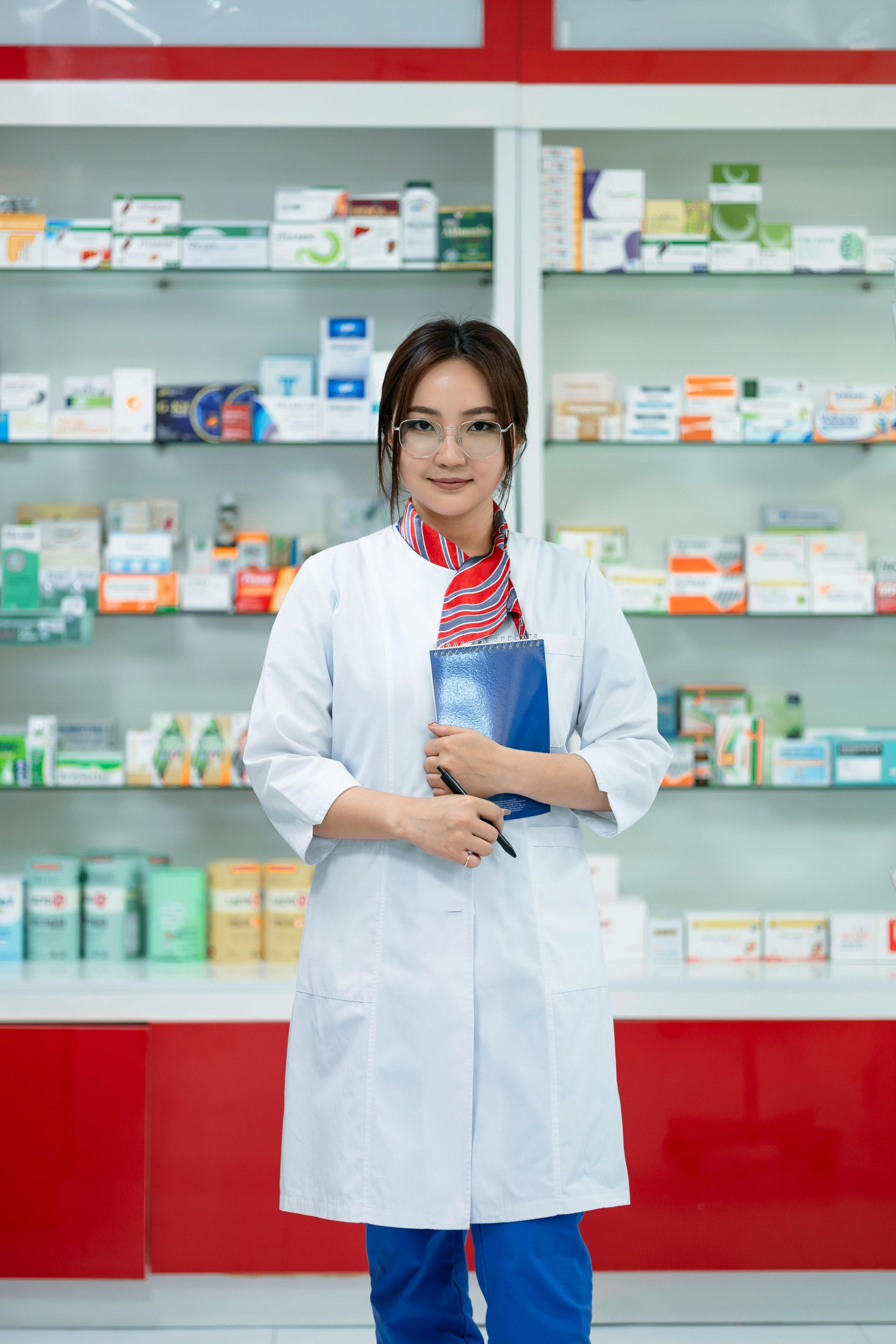 A female pharmacist is standing in front of a shelf in a pharmacy holding a clipboard.