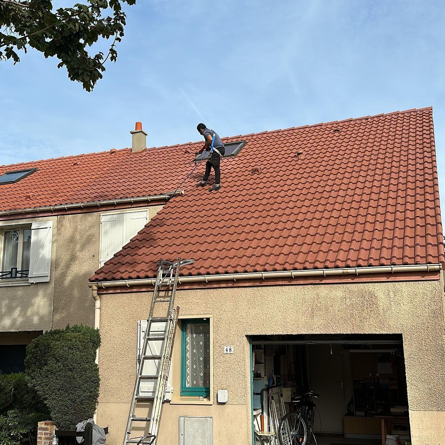 Une personne, perchée sur un toit de tuiles rouges, nettoie une lucarne. Une échelle est appuyée contre la maison.