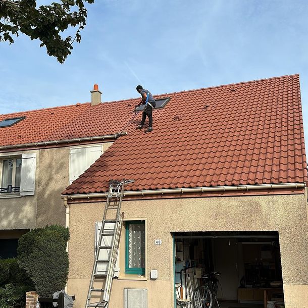 Une personne, perchée sur un toit de tuiles rouges, nettoie une lucarne. Une échelle est appuyée contre la maison.
