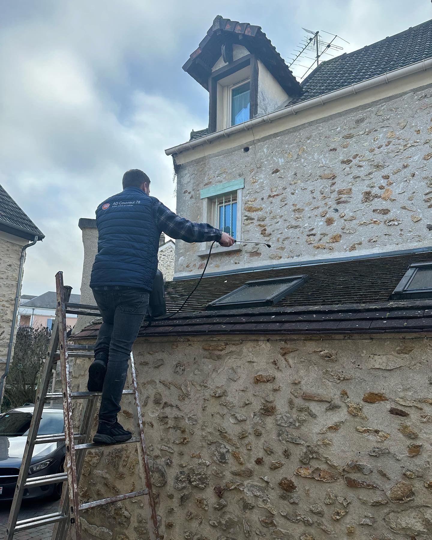 Un homme sur une échelle nettoie le toit au nettoyeur haute pression à côté d'un bâtiment en pierre.