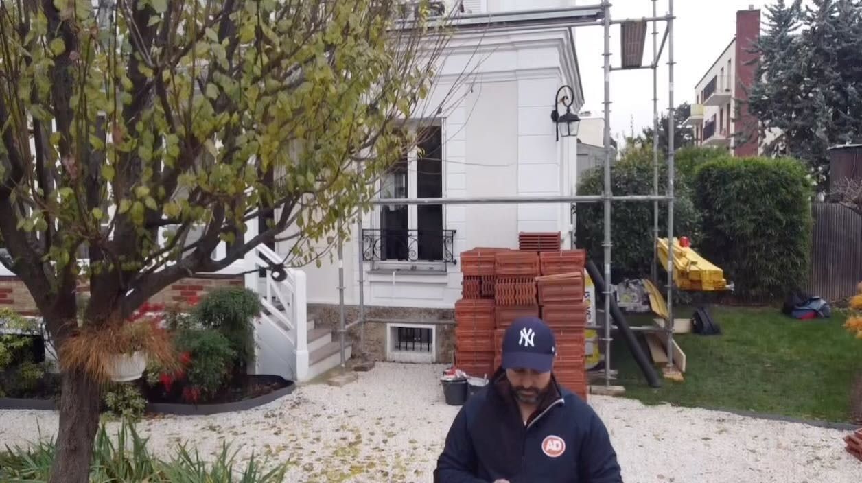 Un homme, perché sur un toit en ardoise, nettoie la gouttière à l'aide d'une longue brosse. Des bâtiments se dressent à l'arrière-plan.