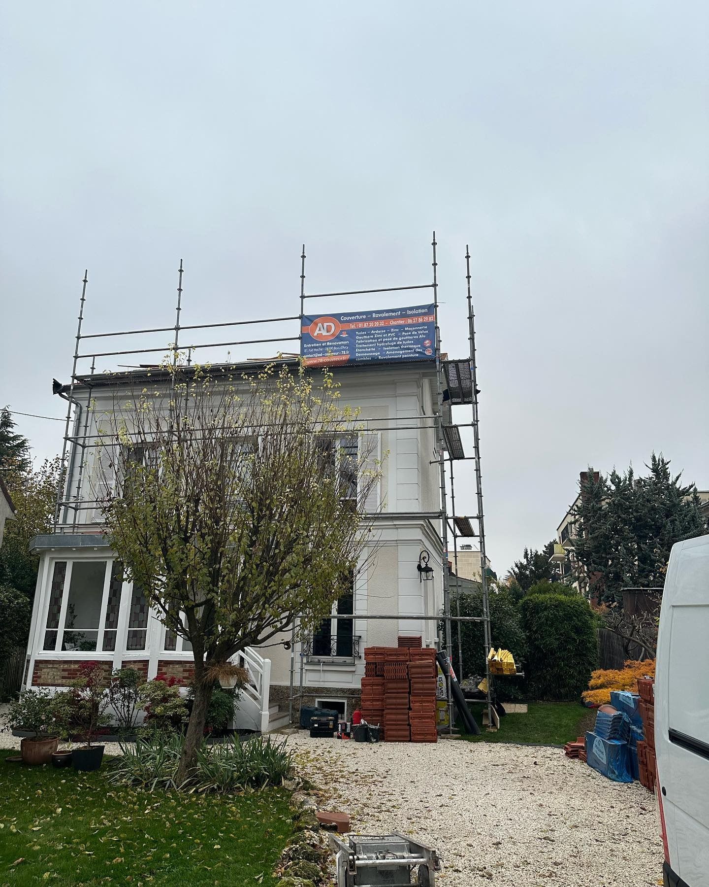 Un camion orange équipé d'une grue soulève des palettes de briques devant une maison.