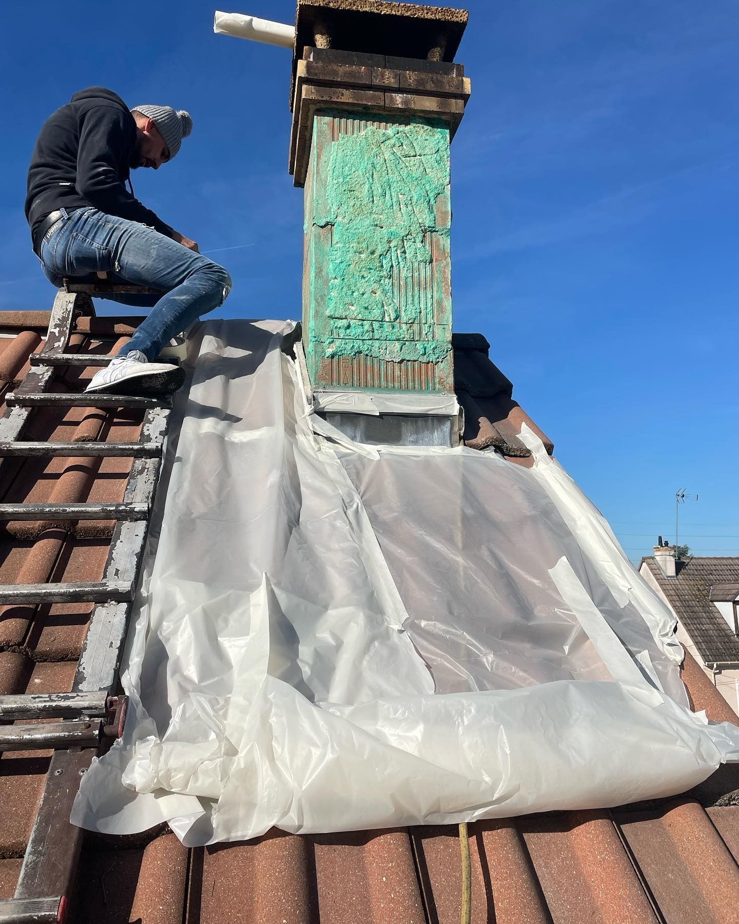 Une personne sur une échelle travaille sur une cheminée. Une bâche en plastique blanc recouvre le toit ; ciel bleu.