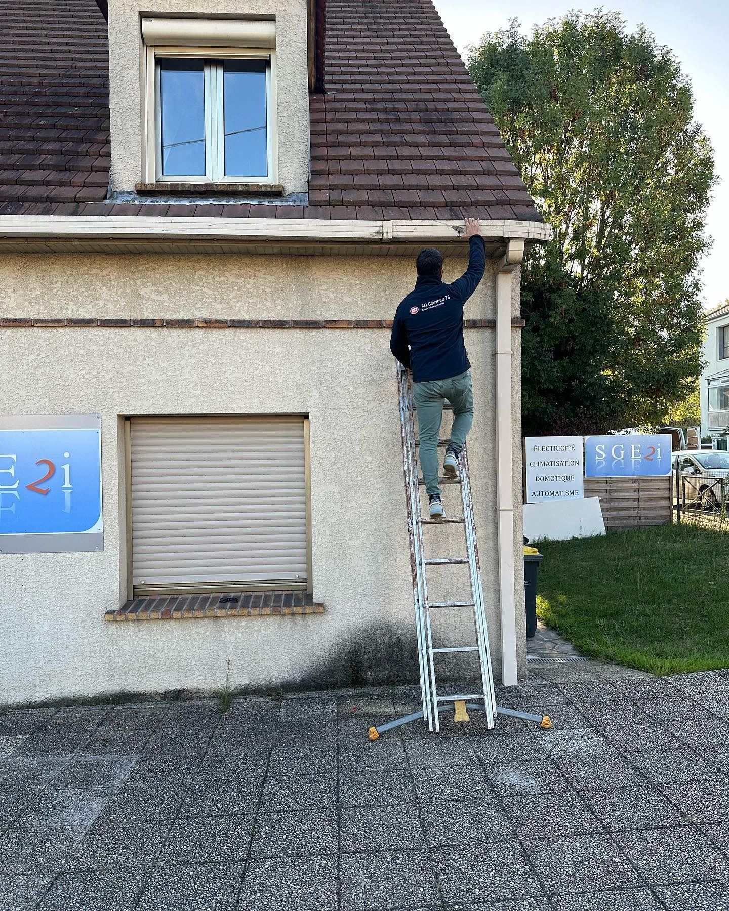 Une personne sur une échelle nettoie la gouttière d'un immeuble. Immeuble beige, toit marron.