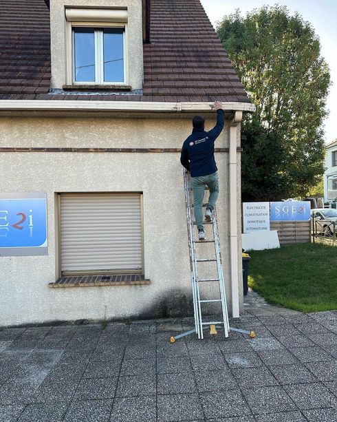 Une personne sur une échelle nettoie la gouttière d'un immeuble. Immeuble beige, toit marron.
