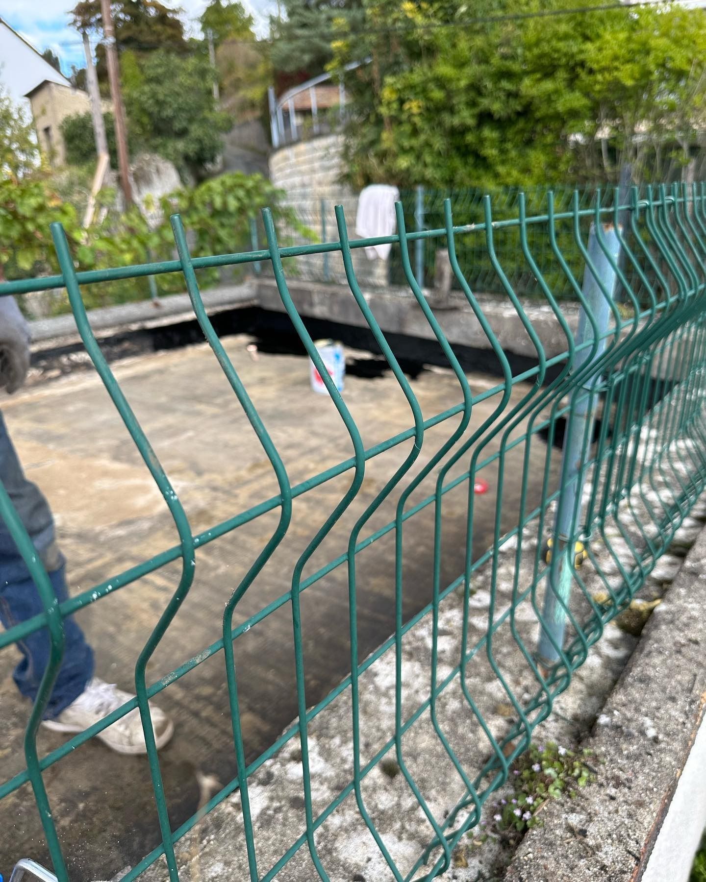 Clôture en fil de fer vert au premier plan ; à l'arrière-plan, une zone en béton avec des taches noires et une personne.