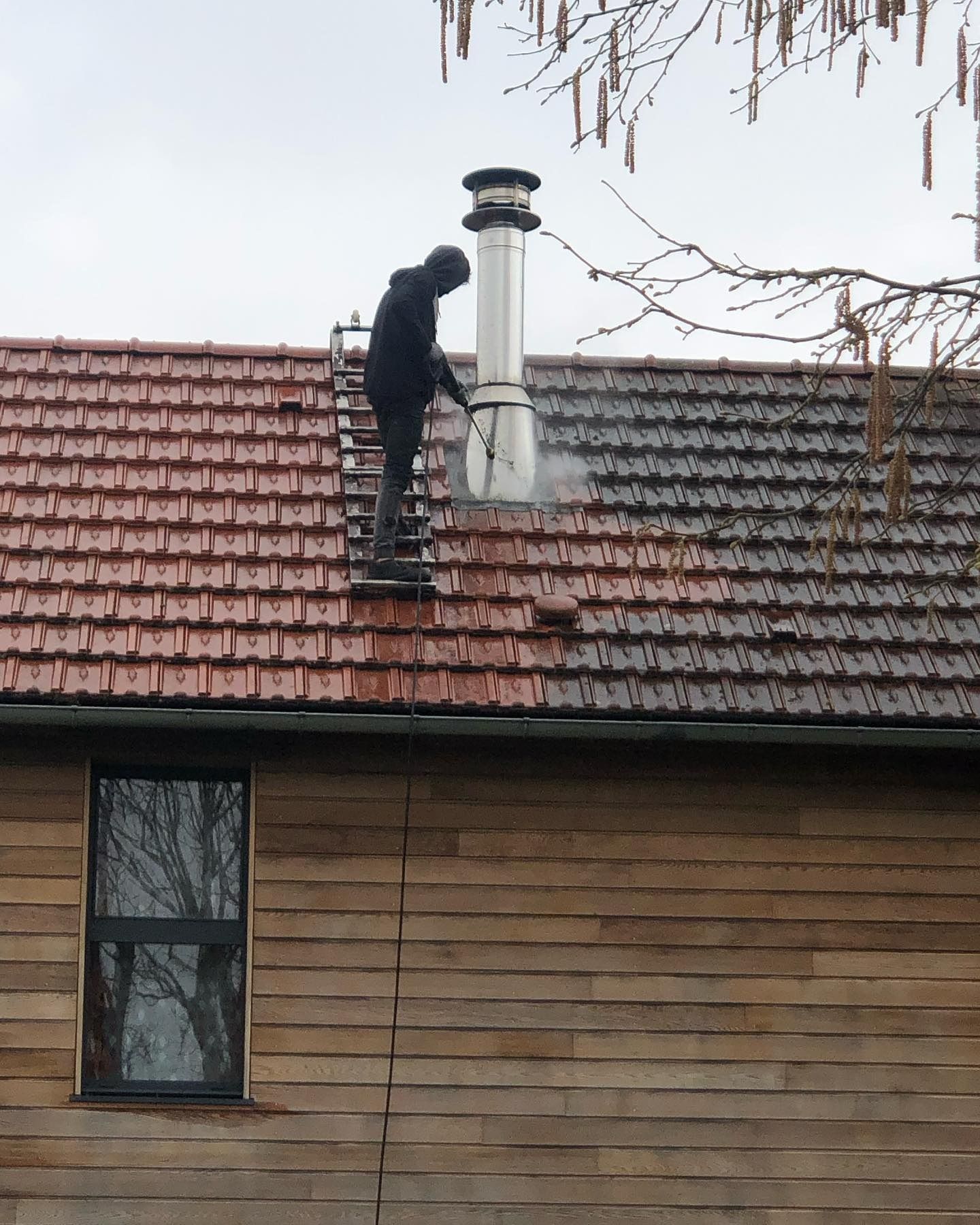 Une personne nettoie au jet d'eau un toit de tuiles rouges, debout sur une échelle. Une cheminée métallique est visible.