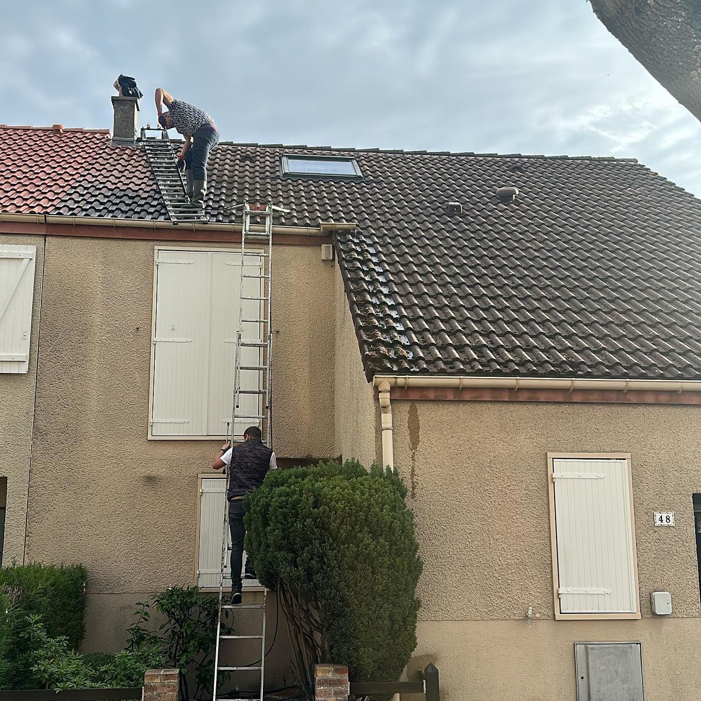Un couvreur nettoie une lucarne sur un toit de tuiles rouges. Une échelle est appuyée contre la maison. Journée ensoleillée.