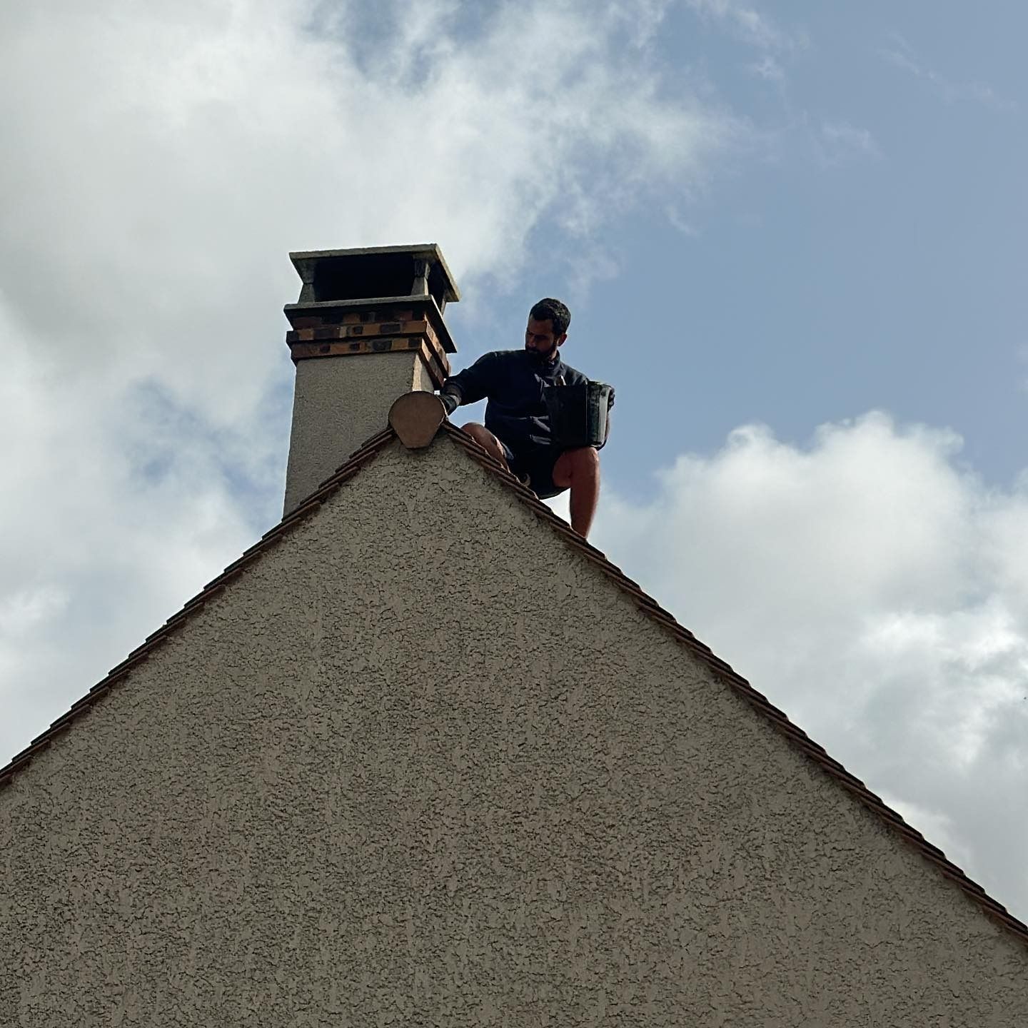 Une personne sur un toit, près d'une cheminée, tenant un seau, sur fond de ciel nuageux.