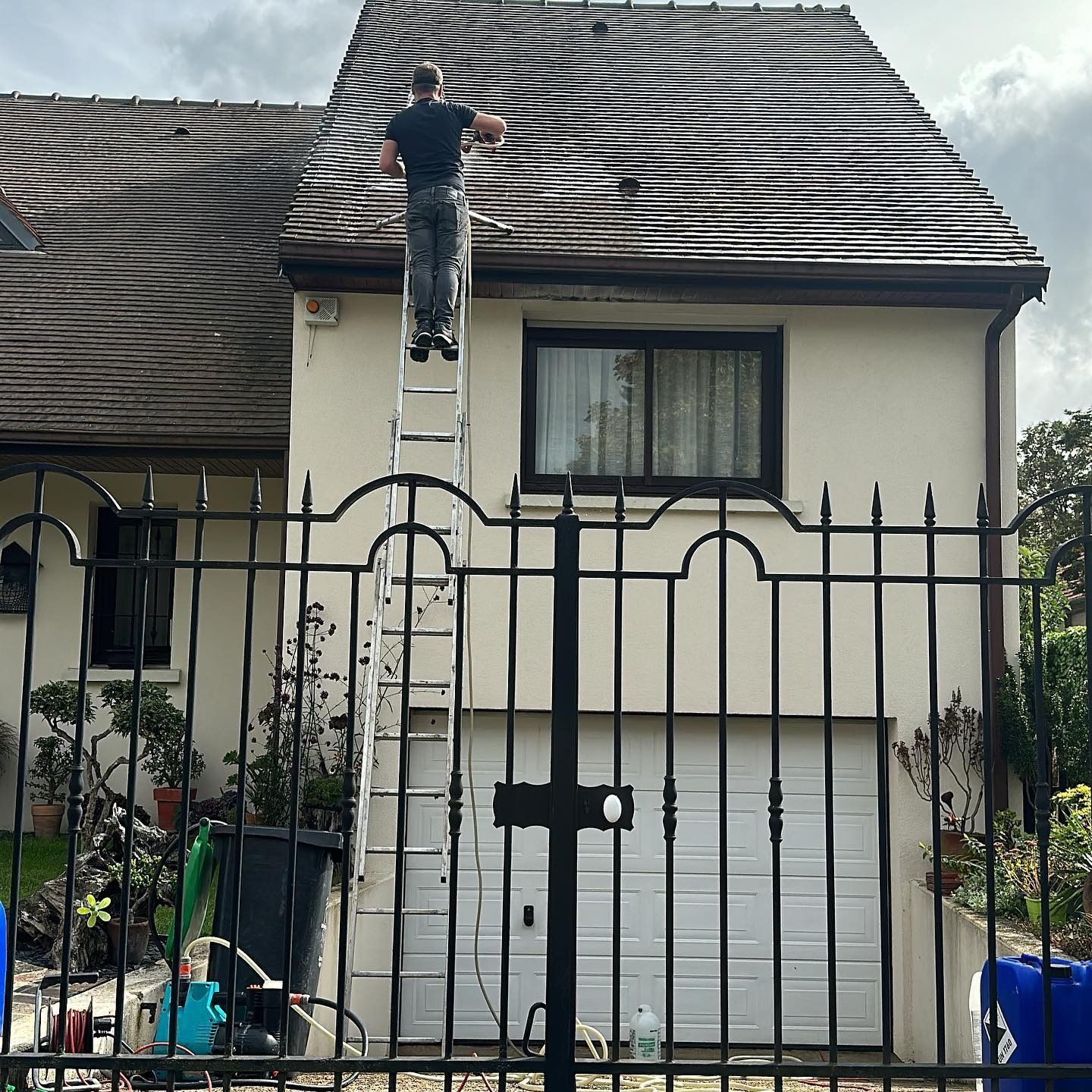 Un couvreur dans le 78, perché sur un toit en ardoise, nettoie la gouttière à l'aide d'une longue brosse.