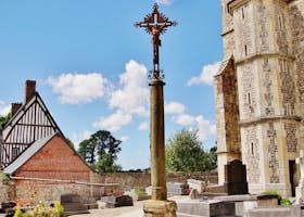 Un grand crucifix se dresse dans un cimetière, à côté d'une église en pierre et d'un bâtiment en briques, sous un ciel bleu.