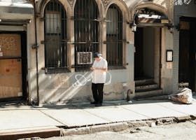Un homme se tient devant un immeuble, regardant vers le bas, tenant quelque chose. L'immeuble possède des fenêtres cintrées, des barreaux métalliques et une porte.