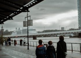 Trois personnes marchent le long d'un sentier au bord de l'eau, surplombant une rivière où se trouvent un grand navire et un pont sous un ciel nuageux.