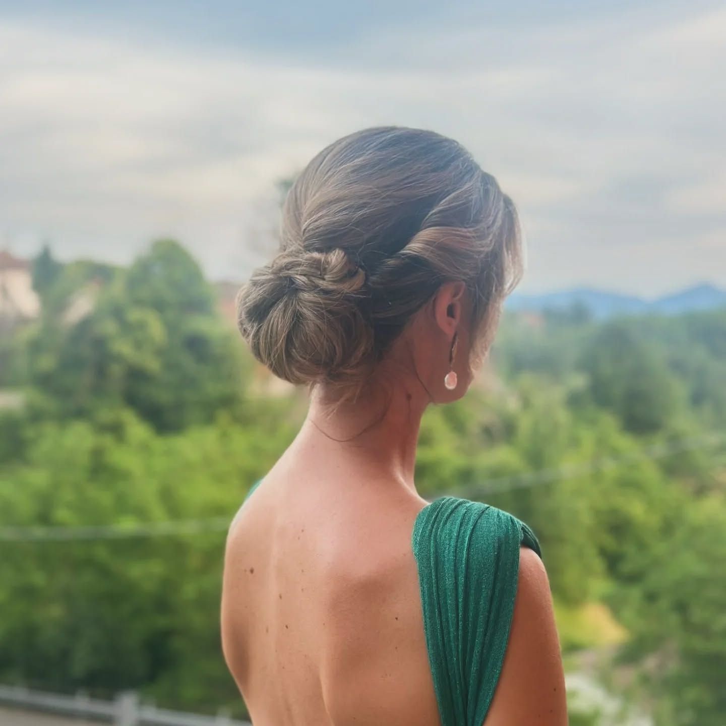 Donna con abito formale verde, di spalle alla telecamera, capelli raccolti, con vista su un paesaggio verde.