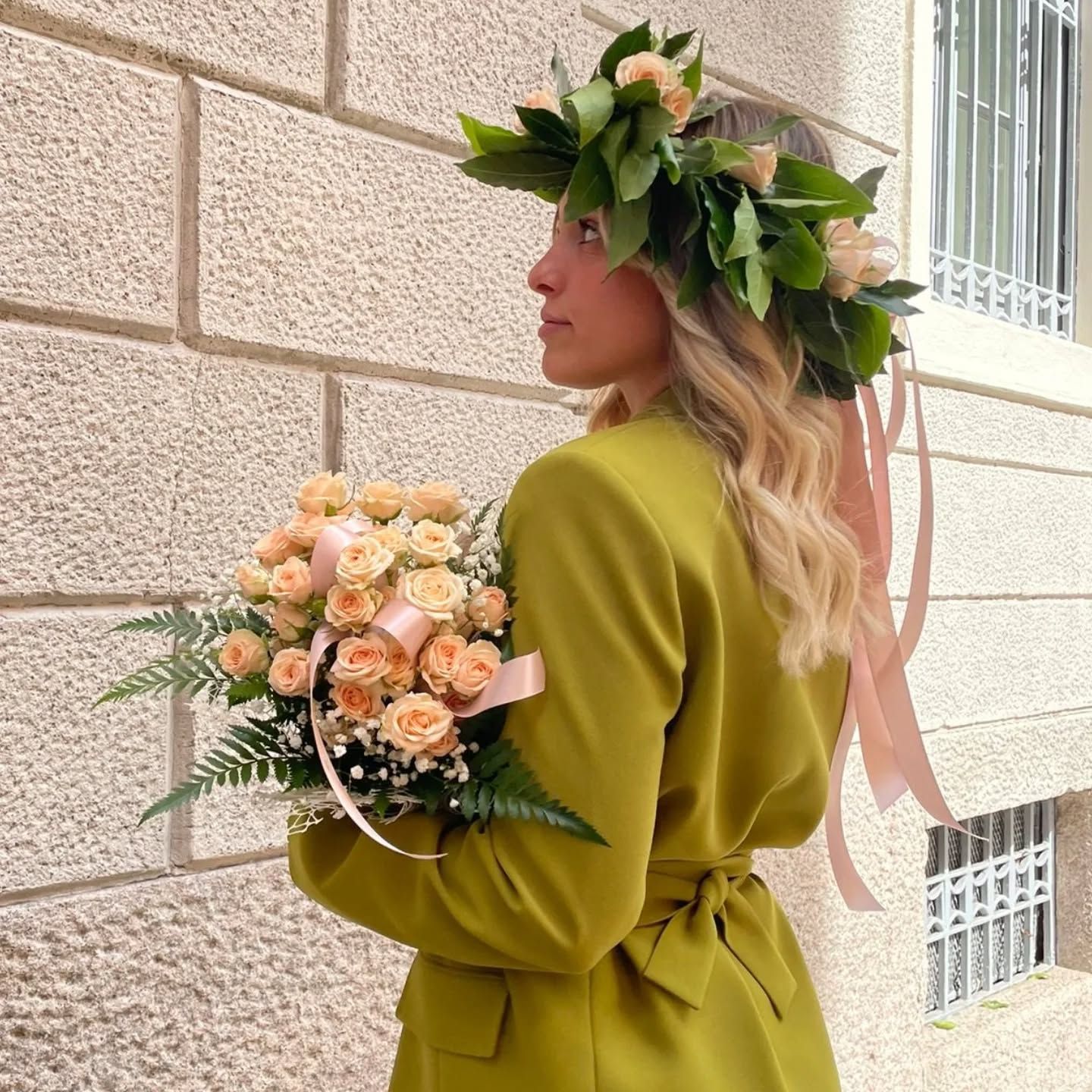 Donna che indossa una corona di fiori e tiene in mano un bouquet, in piedi vicino a un edificio.