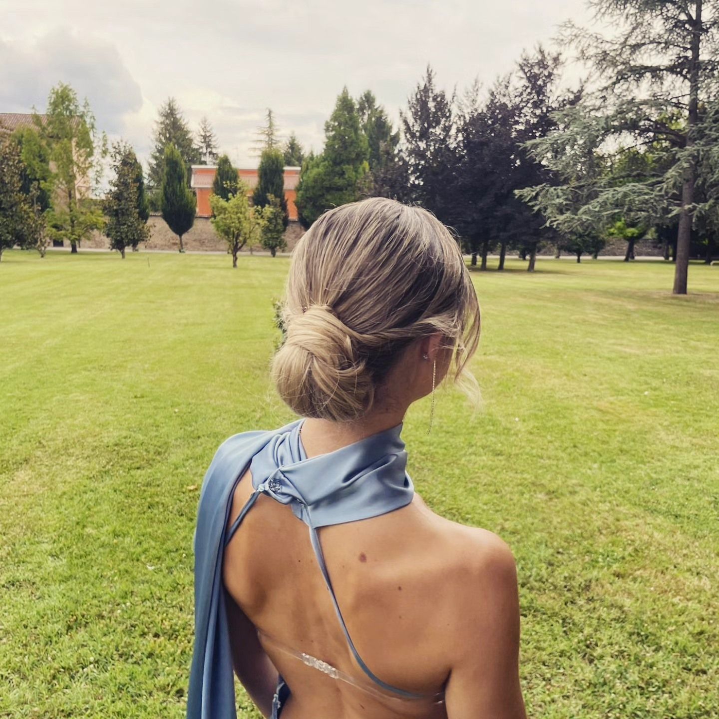 Donna con acconciatura raccolta, che indossa un abito blu, guarda verso gli alberi sul campo erboso.