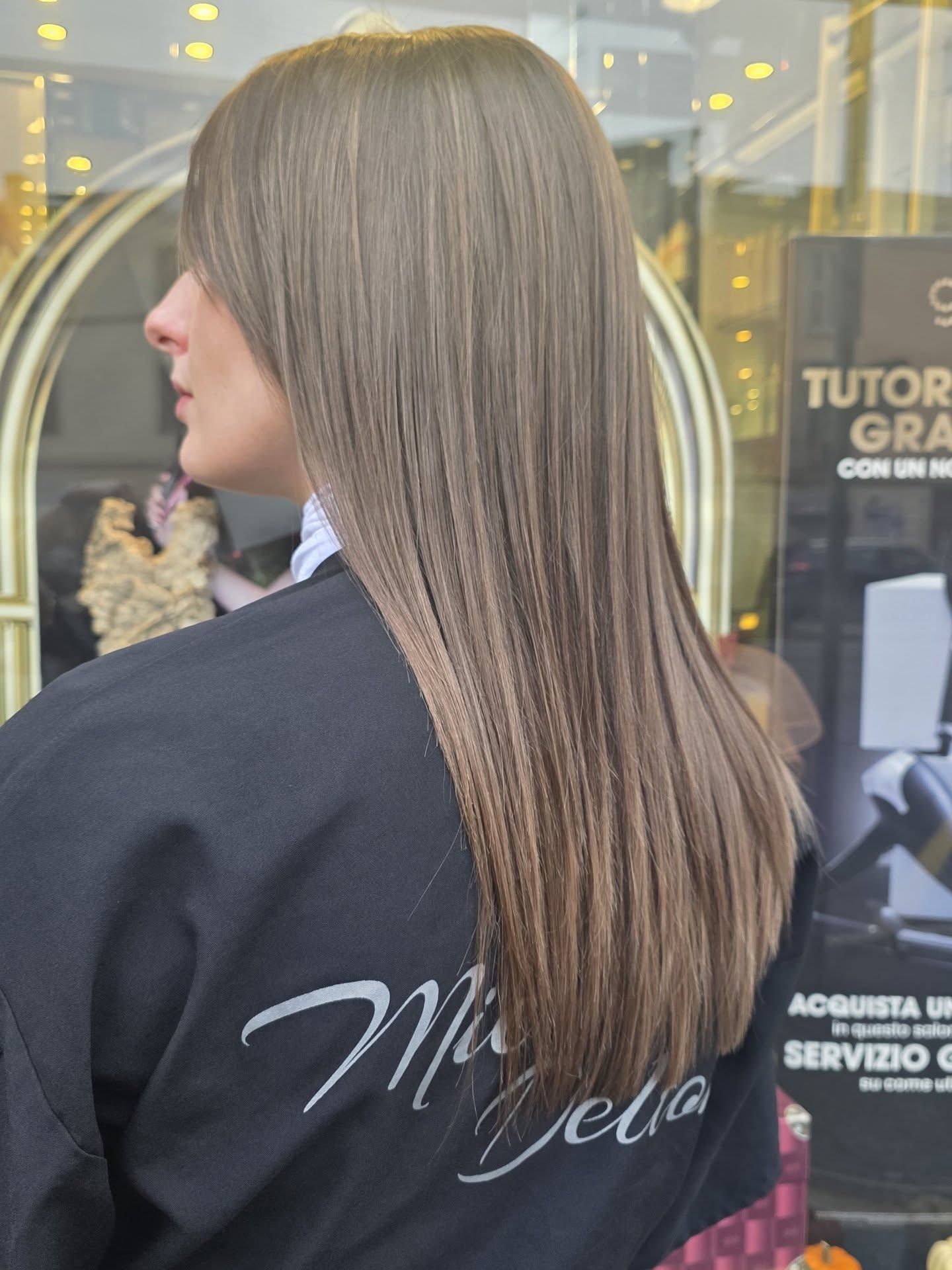 Donna con capelli lisci, castano cenere, in un salone di bellezza. Indossa una mantella nera da salone, davanti alla vetrina di un negozio.